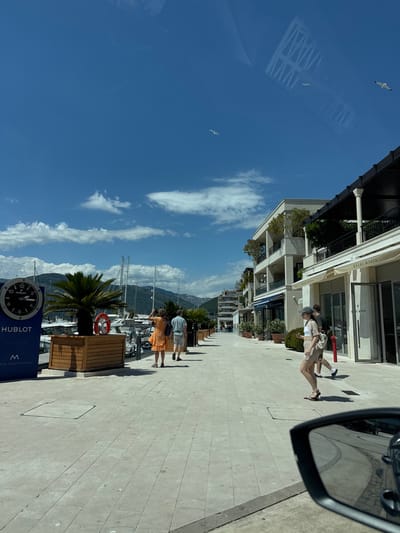 The image depicts a sunny day in Tivat, Montenegro, likely in Porto Montenegro, a luxury marina. The scene is a pedestrian walkway alongside a marina with boats docked. In the foreground, a woman wearing a light-colored outfit and a hat is walking, while a man walks behind her. To the left, a large Hublot clock is visible on a dark blue sign, with a palm tree and a wooden planter box nearby. Several people are walking along the pathway, and there are buildings with balconies and shops lining the right side of the walkway, one of which has a sign that reads "heidi klein." The sky is blue with scattered clouds, and seagulls are flying overhead. The perspective suggests the photo was taken from a vehicle, as a car's side mirror is visible in the lower right corner.