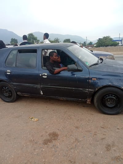 A side view of a dark blue Volkswagen Golf Mk4 hatchback is captured on a dirt road, with a body of water and mountainous terrain in the background. The car's exterior shows signs of wear and tear, with rust and scratches visible along the lower body panels and doors. The car is occupied by at least four individuals. A man in a black shirt is driving, with his right arm resting on the open window frame. Next to him, a woman in a red and blue patterned outfit is seated. In the back seat, two men are visible, one with his head tilted back, and another wearing a white shirt with black trim. One of the men in the back is looking towards the right side of the frame, and another man is standing on top of the car, with his back to the camera. A crumpled plastic bottle lies on the ground near the car's front wheel. The sky is overcast, and the overall lighting suggests it is daytime. The scene appears to be located in Kagini, Nigeria, given the presence of the specific terrain and vegetation.