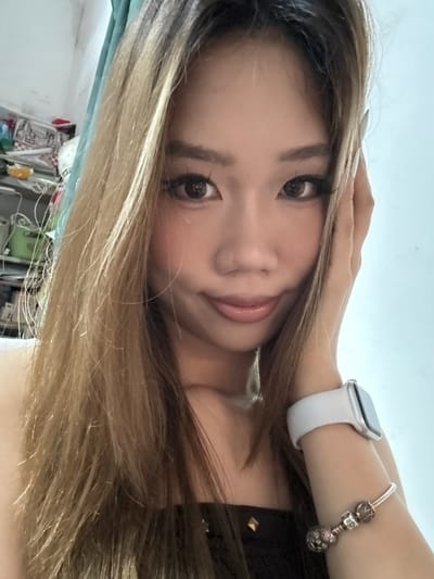 A woman is posing for a selfie. She has long, highlighted hair and fair skin. She is wearing an Apple Watch with a white band and a Pandora bracelet. She is holding her hand near her face. The background is blurry and shows a cluttered area with various objects and items. Based on the available information, the location in the city of Nan'an District, China is undetermined.