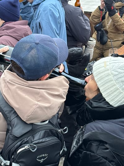 The image captures a close-up of several people seated closely together, possibly in an enclosed space. A woman in a white knit hat appears to be drinking from a dark glass bottle. Nearby, an individual wears a blue baseball cap and another has a tan-colored hood pulled up. An Osprey backpack is visible, as are various winter coats and gloves, suggesting a cold environment. A glass with what appears to be a beverage can be seen among the garments.