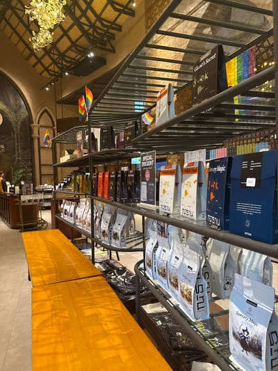 The image shows an indoor setting, likely a retail space like a cafe or shop, with shelves displaying various products. The scene is bright, suggesting it is daytime. The products on display appear to be packaged goods, possibly coffee or tea, with some boxes and bags featuring colorful designs and text in English and what appears to be Thai script. Notable text visible includes "Giant's Secret," "Sorcery Magic," and "Twilight Angel," along with "CHAPTER 2" and "CHAPTER 3". There are also small rainbow-colored flags displayed, which are often associated with LGBTQ+ pride. In the background, architectural elements like arched doorways and a decorative chandelier are visible, hinting at a somewhat traditional or themed interior design. A wooden counter runs along the right side of the image. The overall impression is of a well-stocked retail environment.