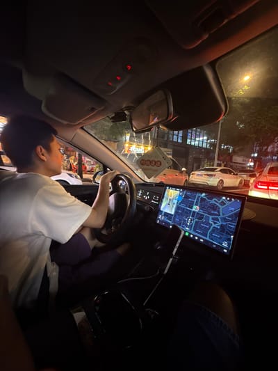 The media file captures a person inside a car at night, likely in Longta, China. The subject is seen from a side view, their face partially obscured as they look forward, their hands on the steering wheel. They are wearing a white t-shirt.
The car's interior is dimly lit, with the dashboard illuminated by a large central touchscreen displaying a navigation map with Chinese text. This screen is the most prominent object in the foreground, alongside the steering wheel and the person's arms. Red indicator lights are visible on the car's overhead console.
Outside the car, the scene suggests a street at night, with car headlights and streetlights creating reflections. Other vehicles are visible on the road, indicating traffic. There are also indications of buildings and illuminated signs, common in an urban environment.
The overall mood is calm, possibly suggesting a commute or a moment of pause in traffic. The time of day is clearly night, due to the darkness outside and the artificial lighting within and around the car. The presence of a navigation map on the screen strongly suggests the activity of driving or being driven. The Chinese text on the screen and the mention of Longta, China, provide specific location context.