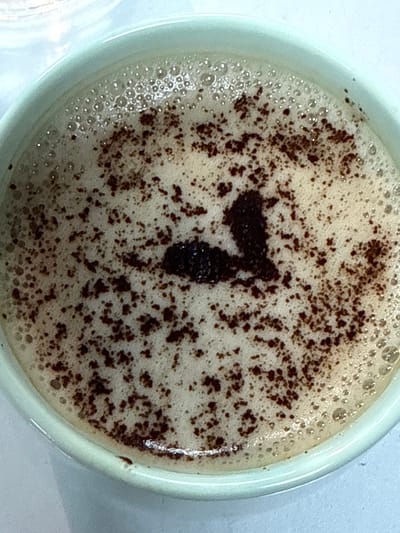 A close-up, overhead shot shows a light green mug filled with a foamy beverage, likely coffee or a latte, dusted with dark brown powder. The powder is sprinkled unevenly across the surface, with a denser clump forming a distinct shape in the center. The foam is bubbly, especially around the edges, and the mug's rim is visible. The beverage is indoors, on a light blue surface.