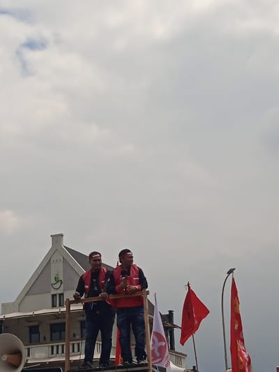 Two men, both wearing red vests and red headbands, are standing on a wooden platform in front of a white building. The building has a gable roof and a green sign on the side. The men are holding microphones and are looking out at the crowd. There are two red flags in the background, one behind the men and one to the right. There is also a white flag with a red symbol on it in the foreground. The sky is cloudy. The location is at Jalan Jendral Sudirman, Yogyakarta, Indonesia (latitude: -7.783, longitude: 110.367). A white loudspeaker is visible on the left side of the frame. The men are speaking into the microphone.
