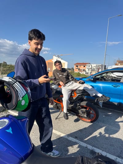 This image captures two young adult males and three vehicles in an outdoor parking area in Mirandela, Portugal, under a clear blue sky with minimal cloud cover.
The male on the left, wearing a dark blue Nike hooded sweatshirt, dark jeans, and light-colored sneakers, stands holding a smartphone in his left hand and a motorcycle helmet with green, white, and red graphic elements in his right hand. He is looking down at his phone with a slight smile. A blue Yamaha MT series motorcycle is partially visible in the immediate foreground, to his left.
The male on the right, wearing a dark brown hooded sweatshirt, light grey sweatpants, and dark athletic shoes, sits casually on an orange and black sports motorcycle, which is resting on its side stand. His left leg is extended forward, and his right leg is bent. He holds a small black object in his right hand and looks towards the camera with a smirk. Behind him, a blue SUV with an "IVECO" logo visible on its rear quarter is parked.
The ground consists of grey asphalt with faded white parking lines. In the background, there is sparse vegetation, an orange-brown residential building, and a multi-story building under construction with exposed concrete frames and red brickwork. A yellow construction crane is visible in the far distance, behind the buildings. A tall street light pole stands on the right side of the frame. Strong sunlight casts distinct shadows across the scene.
