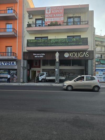 This is an eye-level, medium-shot photograph taken from across a street in Athens, Greece, showing the facades of several buildings. The buildings are multi-story structures with shops and businesses on the ground floor and apartments or offices above. The street in the foreground is paved and has double white lines. A light-colored, four-door hatchback car is driving from right to left in the middle ground.
The building on the left has an orange facade with balconies and windows. A sign that reads "HOTEL" is visible. Below this, a shop displays "LYDIA" in large blue letters. Further down, a shop front shows various battery brands, including "VARTA" and "SINERGY." A man in a white shirt and dark pants is walking on the sidewalk in front of this shop.
The central building is cream-colored with pink trim. On the upper floor, a banner advertises "ΣΧΟΛΕΣ ΧΟΡΟΥ ROSE KARRA" with a phone number. Balconies with railings and plants are visible on this level. The next floor down has green camouflage netting over a balcony. On the ground floor, a dark wood sign reads "KOLIGAS WOOD ESSENCE EST. 1973." Below this, a red graphic with a woman in a dress is visible. A white truck with a closed cargo bed is parked in front of a closed storefront with metal roll-down shutters. A set of stairs leads up to an entrance.
To the right, another building has balconies and awnings. Signs advertising building materials like "XTROMA TA" and brands such as "YTONG" and "KNAUF" are visible. The sky above is overcast. The overall impression is of a commercial street in an urban environment.