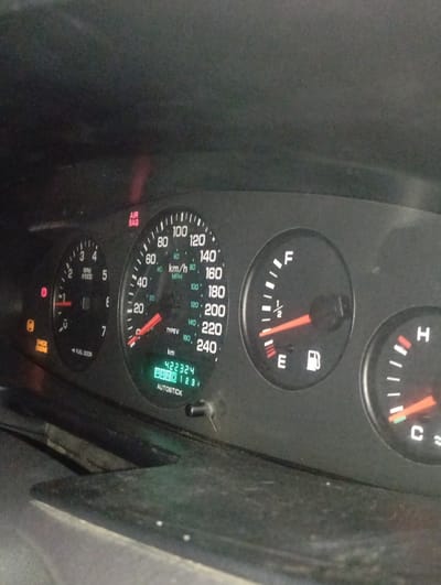 This image depicts an automobile dashboard instrument cluster, located within a vehicle in Kurganinsk, Russia. The scene is viewed from the driver's perspective, displaying a dark gray dashboard surface with visible dust. The speedometer, marked 0-240 km/h (0-150 mph), indicates a speed of approximately 20-30 km/h (15-20 mph). The tachometer shows a needle positioned just above 0 RPM. The fuel gauge needle rests near 'E' (Empty), with the fuel pump icon visible. The temperature gauge needle is at the 'C' (Cold) mark. Several warning indicators are illuminated: a red 'AIR BAG' light, an orange 'CHECK ENGINE' light, and a red circular light with an exclamation mark. The digital odometer displays '422324 km'. Below the odometer, 'P' (Park) is illuminated in green, alongside 'RND 1 2 3' and the text 'AUTOSTICK'. No individuals or specific actions are visible.