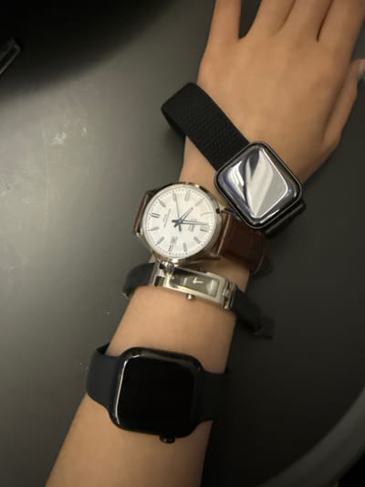 The image shows a person's arm with three watches. The topmost watch is a black smart watch with a square face. Below that is a traditional analog watch with a white face and a brown leather band. The bottom watch is another smart watch, this one black with a square face. The arm is resting on a dark, textured surface, likely a desk or table. The background is dark and out of focus. There is no discernible location in the city of Kommunarka, Russia.