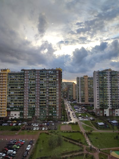 This media file captures an exterior view of a residential area in Murino, Russia. The scene is dominated by several multi-story apartment buildings with a mix of colored facades, primarily brown, yellow, and white, and many windows, some with green balconies. The buildings are arranged along a street lined with parked cars, indicating a moderately populated urban or suburban environment. In the foreground and middle ground, there are patches of green grass, some with pathways and playground-like structures, suggesting a residential complex with common areas.

The sky is filled with dramatic, heavy clouds, with patches of sunlight breaking through, suggesting either dawn or dusk. The weather appears to be overcast, possibly with light rain, as evidenced by what looks like water droplets or streaks on the camera lens. The overall mood is somewhat somber due to the cloudy sky and the muted colors of the buildings, but the light breaking through offers a touch of hope. No visible text is present in the image.