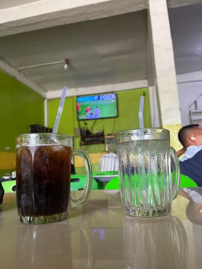 The image depicts a brightly lit interior scene, likely a cafe or restaurant, in Manyang Cut, Indonesia. The setting is casual, with green walls visible in the background and a television screen displaying what appears to be a sports program. The focus of the image is on two large glass mugs with handles, positioned side-by-side.

The mug on the left contains a clear, iced beverage, possibly water or a light-colored soda, with a translucent plastic straw inserted. Condensation covers the outside of this mug, indicating the drink is cold. The mug on the right contains a dark, iced beverage, likely coffee or iced tea, also with a straw. This mug also shows significant condensation. Next to these two mugs, a third, empty glass mug is visible, and its reflection is prominent on the smooth, reflective surface below the mugs.

In the upper background, a person's head and shoulders are visible, with the person looking down. They are wearing a white t-shirt underneath a dark blue shirt. The lighting suggests it could be daytime, but the internal setting makes it difficult to determine the exact time. There are no obvious weather cues. The presence of food and drink items, along with the television, suggests a relaxed atmosphere where people might be enjoying a meal or watching entertainment. The overall impression is one of a casual, everyday moment captured in an Indonesian establishment. A faint, stylized text "REX" is visible on the television screen.