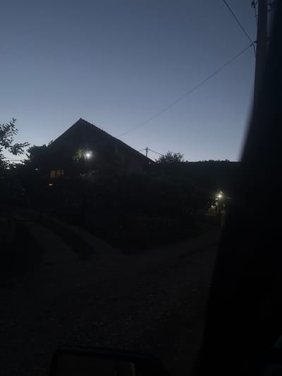 The image depicts a dark, rural scene at dusk in Sevlievo, Bulgaria. A dark silhouette of a house with a steeply pitched roof dominates the center of the frame, partially obscured by dense foliage. A light shines from a window on the left side of the house, indicating occupancy. Another light, possibly a streetlamp, illuminates a patch of ground to the right of the house. A road, composed of gravel and dirt, is visible in the foreground, leading towards the house. Power lines crisscross the sky, a common sight in rural Bulgarian landscapes. The overall atmosphere is tranquil and slightly mysterious, typical of a village at the end of the day.