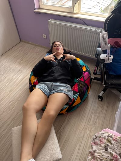 The image captures a single young female individual lying on her back on a colorful bean bag chair within an indoor room, identified as being in Kyustendil, Bulgaria. The subject is dressed in a black zip-up hooded jacket and light blue denim distressed shorts. She has dark hair, dark eye makeup, and dark lipstick, looking directly towards the camera with her mouth slightly agape. Both of her hands are raised, forming a "rock on" gesture with the index and pinky fingers extended and thumbs tucked in. Her left leg is bent at the knee, while her right leg is extended, resting on a white, textured footstool placed on the floor.
The bean bag chair is adorned with a vibrant, multi-colored geometric pattern composed of triangles, lines, and dots in shades of pink, orange, green, blue, and black. The room's flooring consists of light-colored laminate wood. The wall behind the subject is painted a light lavender or purple hue, below a white-framed window that admits natural light and reveals outdoor greenery. A white, horizontally ribbed radiator is mounted on the wall beneath the window, and a white electrical outlet is visible to its left.
To the right of the subject, a black office or gaming chair with white accents on its base and wheels is present. A small white clip-on fan is attached to its right armrest. A blue bag and a reddish fabric item are draped over the back of this chair. In the lower right corner of the frame, a folded pile of textiles featuring a pink and white floral (roses) and script pattern is visible on the floor. A white door frame is partially visible on the left side of the room.