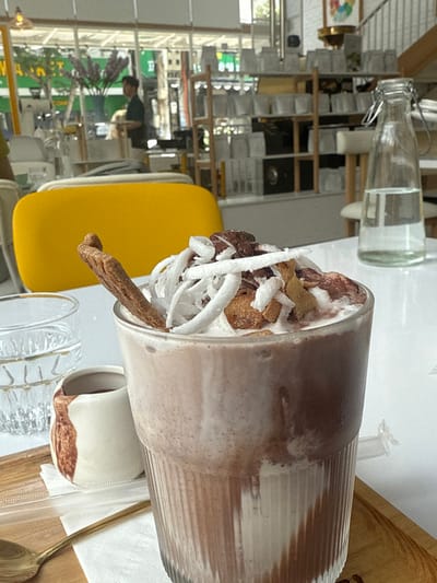 The scene is set indoors at a cafe or restaurant in Ngũ Hành Sơn Ward, Vietnam. The foreground features a tall, ribbed glass holding a layered brown and white beverage topped with whipped cream, white shredded coconut, brown toasted flakes, and a slender, dark brown stick. To the left, a clear glass with water and a small white ceramic pitcher with brown markings rest on a wooden tray, accompanied by a gold-colored spoon and a white napkin. A clear glass bottle with a metal clip-top, partially filled with water, is positioned on the white table to the right. In the background, a yellow chair back is visible. Behind it, a wooden shelving unit displays multiple white-wrapped items. Through a large window, a male individual wearing a dark shirt and light trousers is seen standing, possibly attending to an area, with green foliage and general street elements discernible outside.