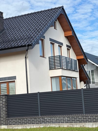 A white stucco house with a dark grey tiled roof is visible. The house has wooden-framed windows, a balcony with a dark grey metal railing, and a dark grey gutter system. A black metal fence with horizontal slats runs across the foreground, supported by brick pillars. The sky is partly cloudy. The image is taken outdoors in the city of Gvardeysk, Russia.