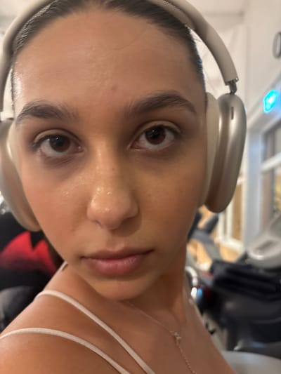 The image is a close-up, forward-facing portrait of a young woman wearing silver over-ear headphones. She has dark hair pulled back and is looking directly at the camera with a neutral expression. Her face is slightly dewy, suggesting she may have been exercising. She is wearing a white camisole with thin straps and a delicate necklace. The background is blurred but appears to be an indoor setting with gym equipment visible, including what looks like treadmills. The lighting is bright and focused on her face. There is no identifiable location in Mönchengladbach, Germany, depicted in this image.