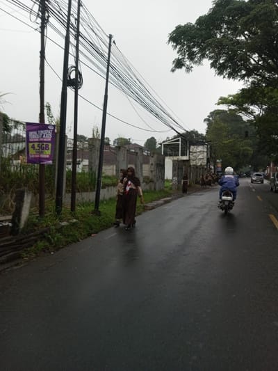The scene is captured in the city of YonPhoogla, Bhutan, on an overcast day with wet asphalt indicating recent precipitation. The image depicts a two-lane road with a yellow dividing line, extending into the distance where multiple vehicles are present. On the left side of the road, three utility poles are positioned, heavily strung with numerous black electrical and communication cables. A purple banner advertising "4.5GB" and "AXIS" is affixed to one of these poles. Adjacent to the road is a narrow strip of grass, followed by a concrete fence, parts of which are covered in graffiti. Beyond the fence, residential structures and dense foliage are visible.
Two individuals, appearing to be young females, walk together on the left edge of the wet road, dressed in matching brown skirts, light-colored tops, and brown headscarves/hijabs. Further down the road, additional individuals in similar attire are also visible near the fence line. A person wearing a white helmet and a blue jacket rides a white scooter away from the viewer on the wet road. Tall green trees line the right side of the road, and an unfinished or renovated building structure with exposed white framing is visible on the left side, further down past the initial fence section. The sky is uniformly grey and overcast.