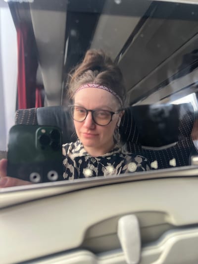 A woman with glasses and a headband is taking a selfie in what appears to be a bus or train, reflected in a window. She is wearing a patterned shirt. The background shows parts of the vehicle's interior, including seats and window frames. The location might be Baltezers, Latvia, given that information, although the image itself doesn't provide explicit details to confirm this location.