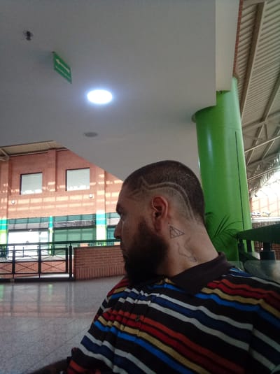 The image shows a man sitting in an indoor public space. The man has a beard, a shaved head with a patterned haircut, and a tattoo on his neck. He is wearing a striped shirt. Behind him is a brick building with windows and a green support column. The ceiling is white with a round light fixture and an emergency exit sign. The floor is tiled. The location is likely Parroquia Guatire, Venezuela.