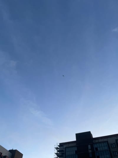 This media file shows a view looking up at a clear blue sky. A small airplane is visible in the distance, flying through the sky. On the right side of the image, the top of a modern building with many windows can be seen. The building's architecture suggests an urban environment. The bright sky indicates it is daytime, likely midday, under fair weather conditions. The composition creates a sense of looking up from ground level between buildings. There is no visible text in the image.