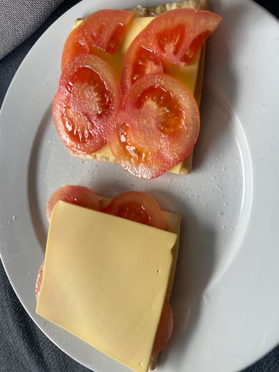 Two open-faced sandwiches are presented on a white plate. The sandwich on the left features a cracker topped with a slice of pale yellow cheese and several slices of fresh, juicy red tomato. The tomato slices are glistening, suggesting they are freshly cut. The sandwich on the right also has a cracker base with a slice of cheese, but only a portion of a tomato slice is visible peeking out from under the cheese. The plate is placed on a dark gray textured surface, possibly a placemat or tablecloth. The lighting suggests an indoor setting, likely during the day. The overall impression is a simple, perhaps homemade, snack. No people are visible in the frame, and no text is present. The setting appears to be a domestic kitchen or dining area.