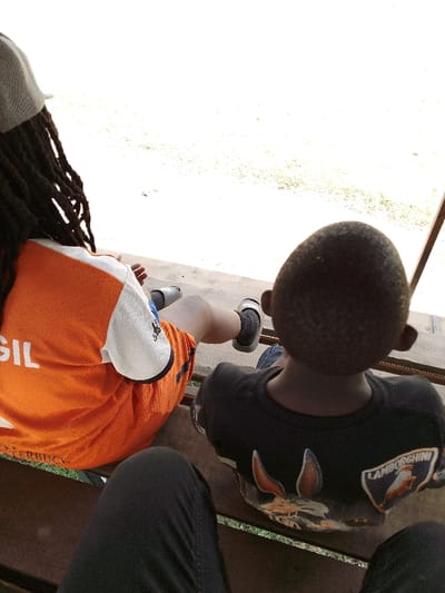 A high-angle, overhead perspective reveals three individuals seated on a dark wooden bench in Nakuru, Kenya. On the left, an individual with dreadlocks wears a grey cap, an orange and white short-sleeved shirt with "GIL" and "ERBUCK" visible, orange shorts, black socks, and grey slides. To their right, a child with short, dark hair wears a dark short-sleeved t-shirt featuring a graphic including a "LAMBORGHINI" shield logo. The camera holder's dark-trousered knees and thighs are visible in the foreground, indicating they are also seated. The background is an overexposed, bright open area with sparse green vegetation visible near the base, and a reddish-brown vertical support structure is present on the far right. All individuals are oriented forward, away from the camera.