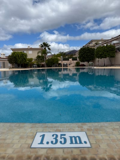 An outdoor swimming pool is depicted under a partly cloudy blue sky in Santiago del Teide, Spain. The foreground features the light brown and beige tiled edge of the pool, with a white sign displaying "1.35 m." in blue text. The pool itself contains clear blue water with light blue mosaic tiles visible beneath the surface, reflecting the sky and surrounding environment. Two stainless steel pool ladders are visible on the far side of the pool, extending into the water. In the background, several multi-story residential buildings with light-colored facades and reddish-brown tiled roofs are present, interspersed with numerous green trees, including a tall palm tree. Distant hills or mountains are visible under the horizon line. No individuals or active subjects are present in the scene.
