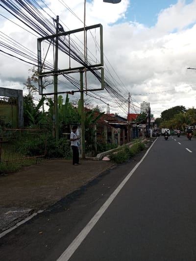This scene is located in the city of Serpang Shalli, Bhutan. The image depicts a street view under a partially cloudy sky.
On the left side of the frame, a man stands on a dirt sidewalk, facing slightly right, looking down at a mobile phone held in both hands. He is wearing a light-colored, short-sleeved collared shirt, dark trousers, and sandals. Immediately behind him, and extending further left, is a dense accumulation of green vegetation, including large-leafed plants and overgrown bushes. A prominent, empty billboard frame, constructed from green metal beams forming two rectangular sections, stands on the left, partially obscuring the background. Numerous black electrical or communication cables are strung overhead between utility poles, many converging on the billboard structure, creating a dense network against the sky. Behind the vegetation and billboard, various single-story buildings with tiled roofs are visible, one featuring a white, minaret-like tower with a conical top in the distance. A rusted metal gate is partially visible on the far left.
The central part of the image is dominated by an asphalt road with a solid white line marking its left edge. Multiple vehicles, including several motorcycles with riders and passengers, and a white van, are traveling away from the viewer. The right side of the road is lined with additional buildings, commercial signage, and mature trees extending into the background. A street light pole is visible on the far right.