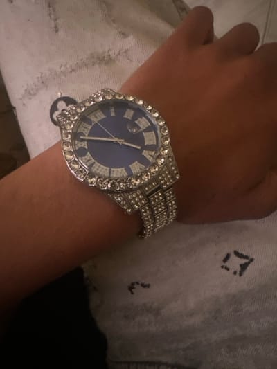 A person's arm is wearing a silver watch with a blue face. The watch has Roman numerals and is encrusted with crystals around the bezel and on the band. The arm is against a light-colored, textured surface. The image does not provide information to determine a location in Macedo de Cavaleiros, Portugal.