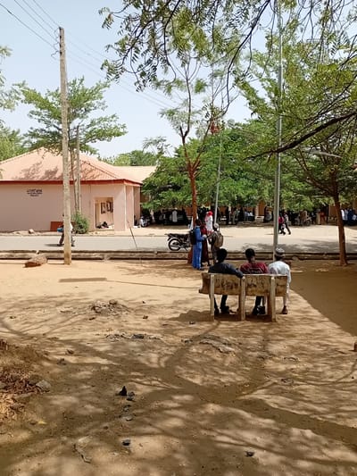 The image shows an outdoor scene in Dabawa, Nigeria. Three people are sitting on a concrete bench under the shade of a tree. A larger group of people is visible in the background, lining a street adjacent to a building with a light pink facade and a red roof. A motorcycle is parked near the group. Overhead power lines are visible between the building and the trees. The ground is dry and sandy.