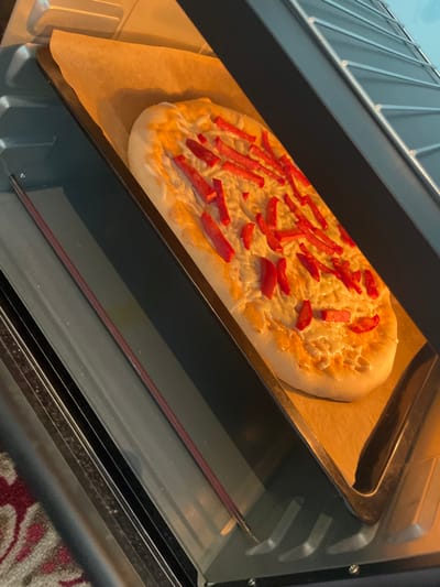 An unbaked flatbread topped with cheese and sliced red peppers is on a baking sheet lined with parchment paper, positioned inside an oven. The oven rack is visible at the top of the image, and the oven door's interior metal framing is also present. The lighting suggests the oven is on, with a warm glow illuminating the flatbread. The scene is indoors, likely a kitchen, but no specific location in Bujum, Kyrgyzstan can be determined from the image.
