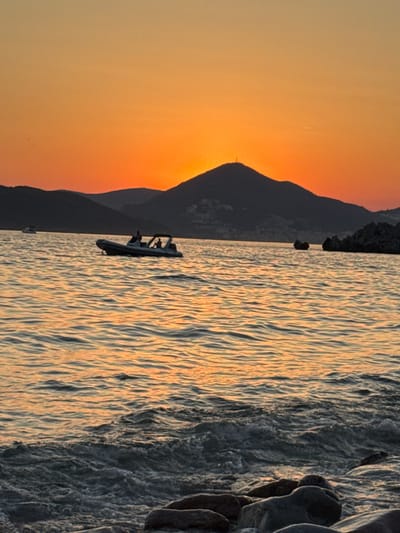 A vertical, medium shot captures a sunset over the sea with a mountainous coastline in Sveti Stefan, Montenegro. The sky is a gradient of deep orange to yellow, with the sun just below the horizon, casting an orange glow on the rippling water. In the foreground, waves crash onto a rocky shore. Further out on the water, a rigid inflatable boat with a canopy and several silhouetted figures is visible. To the left, another boat is a distant speck on the water. The coastline features dark, silhouetted mountains with scattered lights of a town. Two dark, rocky outcrops are visible in the water to the right of the frame.