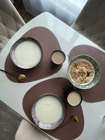 The image depicts a breakfast setting on a white table with brown placemats. Two bowls of white liquid, likely porridge or milk, are visible. One bowl is positioned on the upper left, and another on the lower left. Adjacent to the upper left bowl is a black mug filled with a light brown liquid, possibly coffee or tea, and a brass spoon. To the right of this mug is a decorative bowl filled with puffed rice cereal, also with a brass spoon. A second black mug, containing a similar light brown liquid, is situated on the lower right, below the puffed rice cereal bowl. The scene appears to be indoors, with glimpses of upholstered chairs and a window with blinds in the background. There are no people visible in the image. The location is stated as Podshivalovo, Russia.