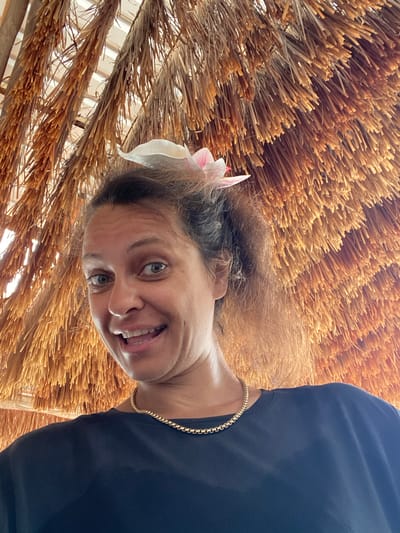 A woman with a flower in her hair and a gold chain necklace smiles at the camera. She is wearing a dark t-shirt. The background consists of a thatched roof made of dried palm fronds. The image appears to be taken in Nusa Penida, Indonesia, likely at a resort or beachside establishment.