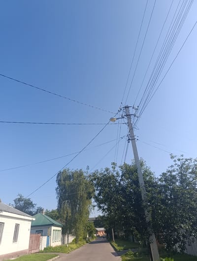 The image captures a street scene under a clear blue sky in Chernihiv, Ukraine. A prominent concrete utility pole with numerous power lines dominates the right side of the frame, with a street lamp attached to it. Trees line the street, with lush green foliage. On the left, a white house with a gray roof and brown trim is visible, adjacent to a smaller building with a green metal roof. The street itself is paved and appears to be a residential road. The overall impression is of a quiet, sunny day in a suburban area.