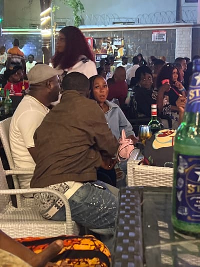 The image captures a lively outdoor social gathering at night, likely at a bar or restaurant in Oba, Nigeria. Several individuals are seated at tables, engaged in conversation and enjoying themselves. In the foreground, two men are prominently featured, one wearing a white t-shirt and a grey cap, and the other in a brown jacket and jeans. They appear to be interacting with each other. A woman with long, dark, curly hair is visible to the left of the men.

The background reveals more people seated at tables, with some holding drinks. There are bottles on the tables, including what appears to be a beer bottle in the lower right corner of the image. The environment is illuminated by artificial lighting, with some decorative lights visible on a structure in the background. A sign with text is present on the wall behind the patrons. The setting suggests a casual and social atmosphere.