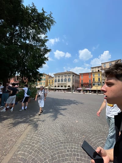 The image captures a sunlit scene in Verona, Italy, showcasing a group of people on a cobblestone square near historical buildings. Dominating the foreground is a young person partially visible in the upper right corner, holding a black smartphone. They are dressed in a light-colored t-shirt. Below, a small group of individuals, seemingly tourists, are walking along the square. One man in a striped shirt and light-colored shorts stands out, holding a camera. The buildings in the background are a mix of yellow, orange, and grey stone, exhibiting distinct architectural styles with windows and balconies. A tree with dense foliage is positioned on the left side of the frame. The sky above is blue with scattered clouds, indicating a clear day.