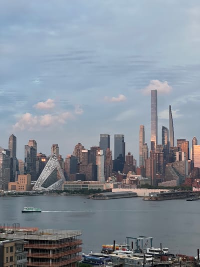 This is a wide-angle, eye-level, exterior shot of the New York City skyline taken from Weehawken, New Jersey, across the Hudson River. The sky is filled with soft, greyish-blue clouds, with a few scattered pinkish-white clouds catching the light. The sun appears to be setting or has recently risen, casting a warm glow on some of the buildings.
In the foreground, the murky grey water of the Hudson River stretches across the bottom half of the image. A green and white ferry boat is traversing the river from left to right, leaving a white wake. In the bottom left of the image, a multi-story building under construction is visible, with its red structural beams and scaffolding exposed. Further to the right, a cluster of boats and barges are docked, including one with the word "Surfside" visible on its side.
The middle ground is dominated by the iconic New York City skyline, featuring a dense collection of skyscrapers of various heights and architectural styles. Notable buildings include the distinctive geometric pyramid-like structure of VIA 57 West, and several very tall, slender residential towers on Billionaires' Row, such as 111 West 57th Street (Steinway Tower) and Central Park Tower, which reflect the sky and clouds. Some buildings have their windows illuminated.
The background consists of more distant buildings and the sky, which extends to the top of the frame. The overall impression is a serene urban landscape, captured during the transitional light of early morning or late evening.