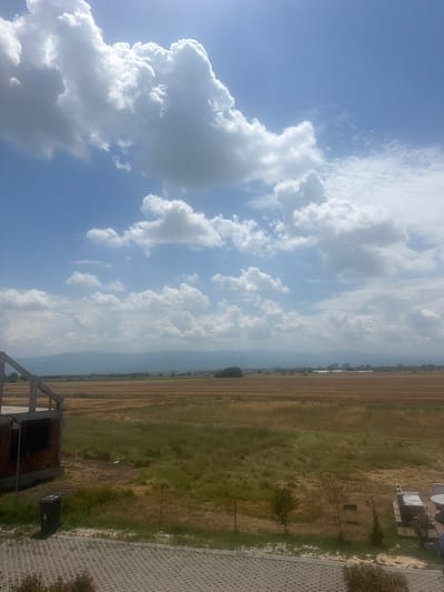 The image presents a wide outdoor landscape in Skutare, Bulgaria, under a clear blue sky populated by numerous large, white cumulus clouds, indicating a bright, sunny daytime environment. In the immediate foreground, a grey interlocking paver patio extends across the bottom, bordered by a strip of dry grass and a few small, sparse shrubs. To the left, a partially constructed building with exposed concrete framing and red brick walls is visible, adjacent to a black waste bin marked with the number "4." To the right, a segment of an outdoor seating area featuring a covered table and chairs is partially visible. The midground consists of extensive, flat agricultural fields displaying a mixed texture of golden-brown stubble from harvested crops and patches of taller, lighter green grasses. The distant background reveals a long, hazy mountain range beneath a band of faint urban structures. No people, animals, or active interactions are present in the scene.