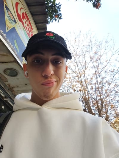 A young adult stands outside with a white hoodie and a black cap embroidered with a red rose. Wireless earbuds are in both ears. Behind him is a building with signage in Arabic and English and a bare tree with a few leaves. A second tree with oranges is visible overhead. The individual looks directly at the camera with a slight smile.
