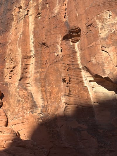 The image shows a rock wall, likely sandstone, with various petroglyphs carved into the surface. The rock is reddish-brown, and there are areas of light and shadow. The petroglyphs are faint and appear as simple line drawings. The environment suggests an outdoor setting, possibly in a desert or canyon area. Based on the location provided, this is likely to be a petroglyph site in the Moapa Valley, United States.