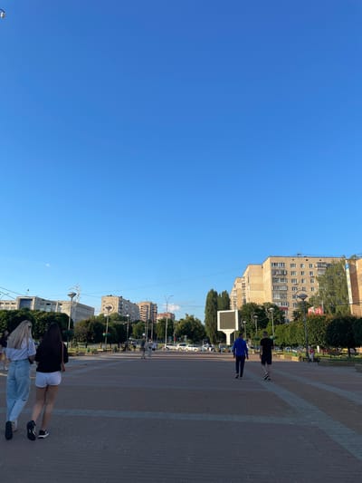 The image shows a wide pedestrian area in Brovary, Ukraine, paved with brickwork. On the left, two young women are walking away from the camera. The woman on the left has blonde hair and is wearing light blue jeans and a white t-shirt. The woman on the right has dark hair and is wearing white shorts and a black t-shirt. Further in the distance, several people are walking and sitting on benches. On the right side of the pedestrian area, two men are walking away from the camera, one in a blue hoodie and dark pants, the other in a black t-shirt and shorts. In the background, there are multi-story apartment buildings and trees. A large, rectangular digital billboard is visible in the middle ground. The sky is clear and blue.