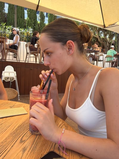A young woman with light brown hair pulled into a bun is seated at a wooden table at an outdoor establishment, possibly a restaurant or cafe. She is wearing a white tank top and a delicate silver necklace, and a pink bracelet is visible on her wrist. She is drinking a red beverage with ice and a garnish from a glass using two black straws. The table also holds a rectangular object, possibly a napkin or a small book.
In the background, other patrons are visible seated at tables under sun umbrellas. The setting appears to be in Cala D'or, Spain, suggested by the presence of Mediterranean vegetation, architectural details, and a general outdoor atmosphere. The day is sunny, as indicated by the brightness and shadows. A white highchair is positioned near the table.