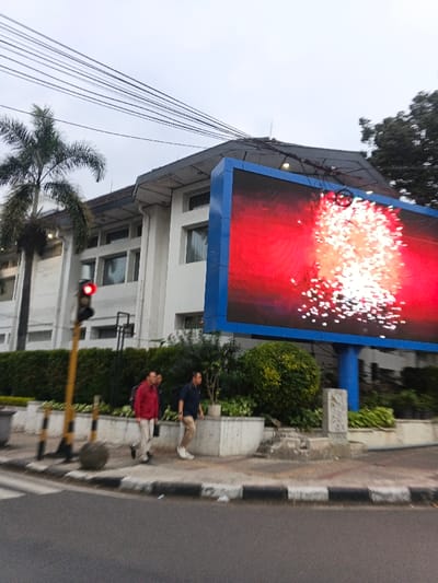 This image captures a daytime urban scene in Bandung City, Indonesia, beneath an overcast sky. In the foreground, an asphalt road with a visible crosswalk and a black and white striped curb transitions to a concrete sidewalk on the right. A yellow-poled traffic light with an illuminated red signal is positioned on the left. Three adult males are walking from left to right on the sidewalk; one is wearing a red long-sleeved shirt, another is partially obscured in a dark shirt, and the third is wearing a dark blue polo shirt and light-colored pants, carrying a dark backpack. A spherical grey concrete object rests on the sidewalk near the traffic light. A low concrete planter, containing green foliage and a white rectangular stone marker, separates the sidewalk from a large, blue-framed LED digital billboard on the right. The billboard displays a vibrant red background with numerous scattered white light patterns, resembling fireworks. Behind the billboard and to its left, a multi-story white building with a dark roof and multiple windows is visible. A tall palm tree stands on the far left, and a dense green tree canopy is visible on the far right. Multiple black utility lines crisscross the upper portion of the frame against the sky.