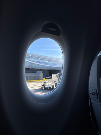 View from an airplane window looking out at the airport terminal at Charles de Gaulle Airport in Le Mesnil-Amelot, France. A modern glass-walled terminal building is visible, along with airport vehicles and equipment on the tarmac. The sky is blue. The view is framed by the airplane cabin interior, including a USB charging port.