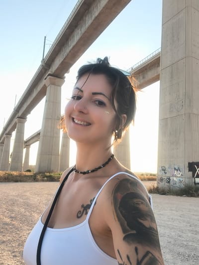 An outdoor, back-lit portrait captures a young woman smiling at the camera, positioned in the foreground against a backdrop dominated by a multi-arched concrete railway viaduct located in the Mate Yehuda Regional Council, Israel. The woman has short, dark hair, a septum piercing, a left nostril piercing, and glitter makeup on her left cheekbone. She wears a white spaghetti-strap tank top, a dark beaded choker necklace, and has sunglasses resting on her head. Visible tattoos include a small, dark lizard on her upper left chest and a large, dark, detailed arm sleeve tattoo on her right shoulder/bicep. A black strap, likely from a bag, crosses her left shoulder. The background features a large railway viaduct, supported by numerous tall, square concrete pillars, extending horizontally across the frame. Another section of a similar viaduct is visible further in the distance to the left. The ground is unpaved earth with sparse, dry vegetation. Graffiti is visible on the lower section of the concrete pillar on the far right. The scene is illuminated by strong backlighting from a low sun, creating a bright halo around the woman's head and hair.