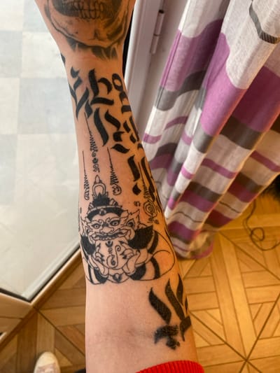 An overhead close-up image shows the tattooed left forearm of an individual in Kunje, Montenegro. The arm features several black ink tattoos: a faded skull on the upper arm, multiple lines of calligraphic script, and a prominent central design depicting a crowned, horned figure with wide eyes and bared teeth, surrounded by smaller symbolic lines. Towards the wrist, a stylized black tattoo resembling flame-like shapes is partially visible, with a red knitted cuff obscuring the lower forearm. The background consists of a light-colored window or door frame and a light wooden floor with a geometric pattern on the left, and a vertically striped curtain with white, purple, and gray bands on the right.