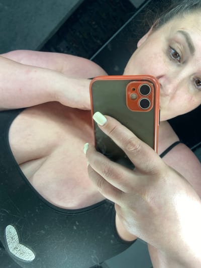 A woman is taking a selfie in Rozkvit, Ukraine. She is holding a smartphone in her right hand, with her left arm resting on her chest. The woman is wearing a black tank top with a white heart logo. Her nails are painted a light green color. The background is dark and appears to be a mirror.