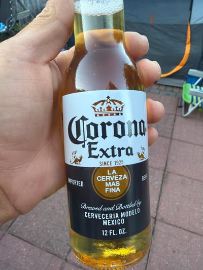 A hand holds a glass bottle of Corona Extra beer. The label on the bottle is white with black text and a crown logo. The text on the label reads "Corona Extra", "SINCE 1925", "LA CERVEZA MAS FINA", "IMPORTED", "Brewed and Bottled by CERVECERIA MODELO MEXICO", and "12 FL. OZ.". The beer is golden in color and has a foamy head. In the background, there is a blurry image of a tent and chairs. The ground appears to be paved with bricks. The image was taken in Riverhead, United States of America.