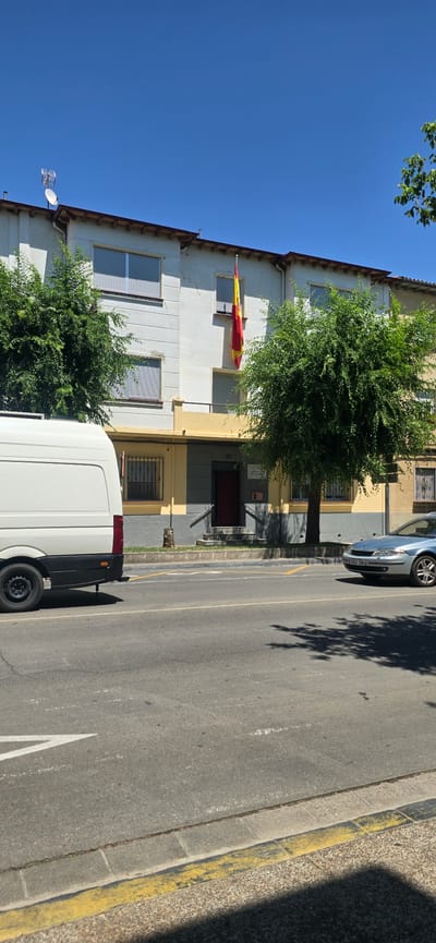 The image shows a building facade on a sunny day, with a Spanish flag hanging from a window. The building is painted in white and yellow, with several windows visible. A tree with lush green foliage stands in front of the building. A white van is parked on the street next to the building, and a silver car is parked further down the street. The street is paved and has a yellow curb. The sky is clear and blue, suggesting good weather. There are shadows cast by the tree and the buildings, indicating it is daytime. The scene appears to be a residential or commercial area. A small sign with the number "37" is visible on the building. The overall impression is a peaceful, everyday scene.