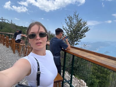 This is a selfie taken outdoors by a woman with long brown hair, wearing sunglasses and a white t-shirt. She is sitting at a wooden table. Behind her, a man wearing a dark blue t-shirt and a dark bag slung across his chest is visible, holding a phone. Further in the background, there are more people sitting at tables and chairs, with wooden fencing and greenery. The scene appears to be a scenic overlook, with a view of a valley and a distant body of water under a blue sky with scattered white clouds. A roller coaster track is visible in the far background. The overall impression is a relaxed, scenic outing on a sunny day.